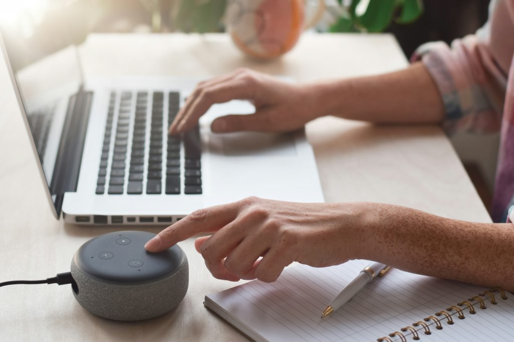 Woman sitting at desk using laptop computer and pressing button on virtual assistant smart speaker